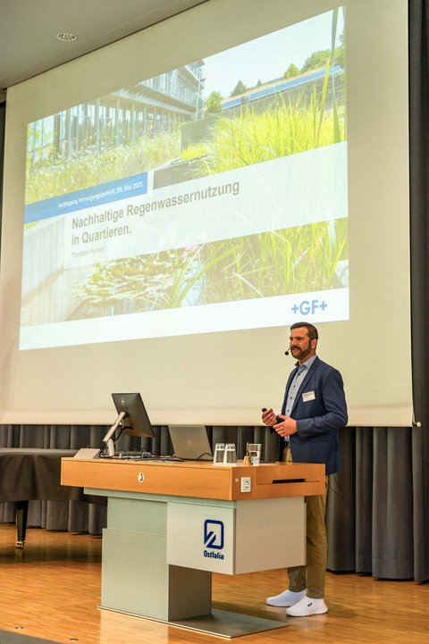 Aufnahme eines Speakers auf der Bühne in der Aula der Ostfalia Hochschule. Im Hintergrund ist die Präsentation zum Thema Nachhaltige Regenwassernutzung in Quartieren. (öffnet Vergrößerung des Bildes)