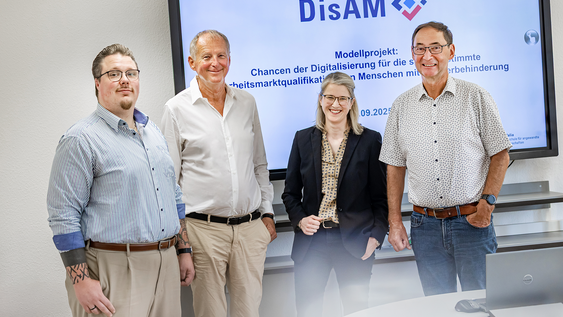 Einige Personen aus dem Team des Modellprojekts stehen vor einem großen Monitor, auf dem das Logo des Projekts zu sehen ist.