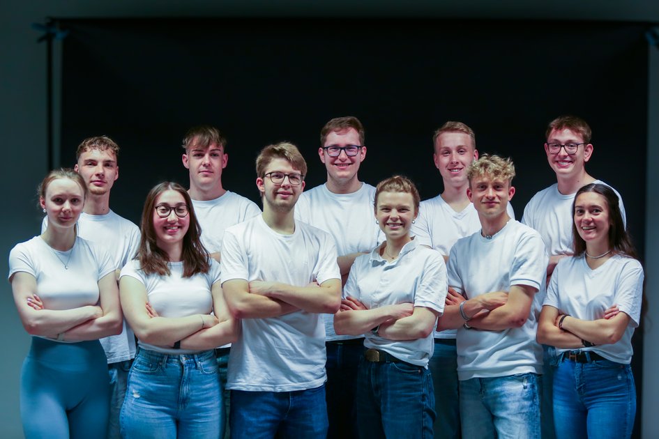 Eine Gruppe von Studierenden steht in weißen Oberteilen mit verschränkten Armen vor einem dunklen Hintergrund