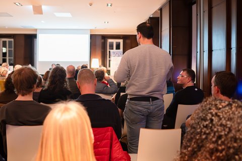 Ein stehender Mann aus dem Publikum ist von hinten zu sehen, wie er eine Frage an die Teilnehmer:innen der Podiumsdiskussion stellt. (öffnet Vergrößerung des Bildes)
