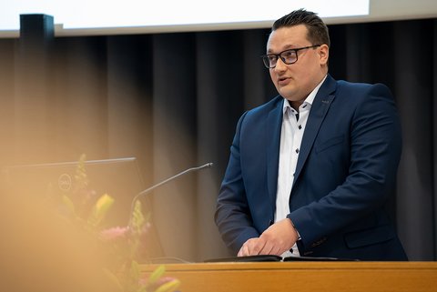 Nils Fründ bei seinem Alumni-Grußwort am Rednerpult. Er trägt einen blauen Anzug und ein weißes Hemd. (öffnet Vergrößerung des Bildes)