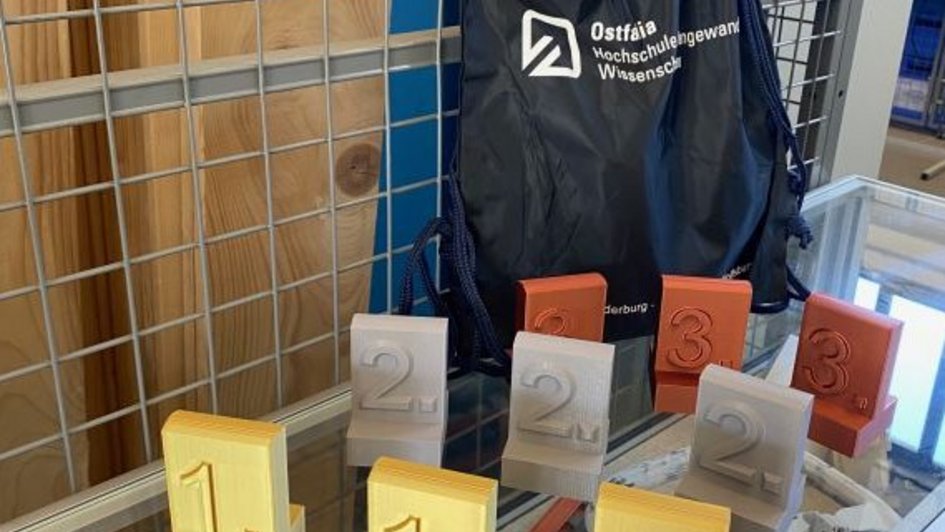 Auf dem Bild sind Pokale für einen ersten, zweiten und dritten Platz zu sehen. Die Pokale wurden im 3D-Drucker gefertigt. Hinter den Pokalen steht ein kleiner Rucksack mit Ostfalia-Schritzug. (öffnet Vergrößerung des Bildes)