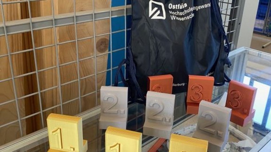 Auf dem Bild sind Pokale für einen ersten, zweiten und dritten Platz zu sehen. Die Pokale wurden im 3D-Drucker gefertigt. Hinter den Pokalen steht ein kleiner Rucksack mit Ostfalia-Schritzug. (öffnet Vergrößerung des Bildes)