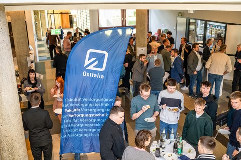 Aufnahme von oben außerhalb der Aula. Die Zuschauer stehen versammelt in Gruppen, unterhalten sich, essen und trinken. Im Vordergrund ist die Flagge der Ostfalia Hochschule. (öffnet Vergrößerung des Bildes)