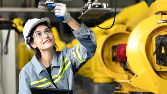 Eine Studentin mit Sicherheitshelm steht an einer großen Maschine. Sie schaut nach oben und prüft die Maschine.