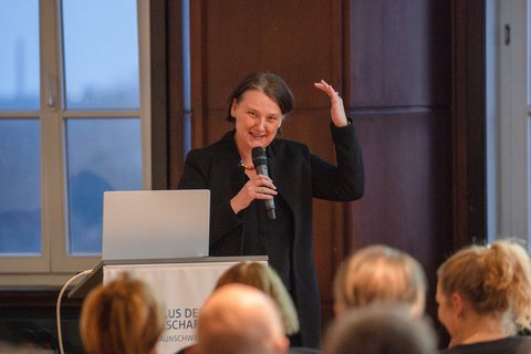 Prof. Dr. Britta Wrede steht vorne am Pult, halt ein Mikrofon in der einen Hand, hebt die andere Hand leicht über den Kopf und spricht zum Publikum. (öffnet Vergrößerung des Bildes)
