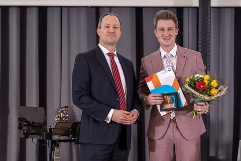 Sören Damerau und Leander Becker im hellen Anzug. Leander Becker hält den PKF Fasselt-Award, eine bronzefarbene Münze im Rahmen und einen Bluenstrauß. Die Abgebildeten lächeln in die Kamera. (öffnet Vergrößerung des Bildes)