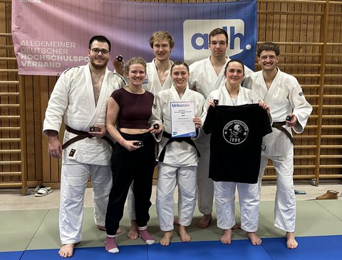 Sieben Judoka posieren nach der Deutschen Hochschulmeisterschaft vor einem adh-Banner, halten ihre Bronzemedaillen und eine Urkunde und zeigen ein schwarzes Judo-Teamshirt. (öffnet Vergrößerung des Bildes)
