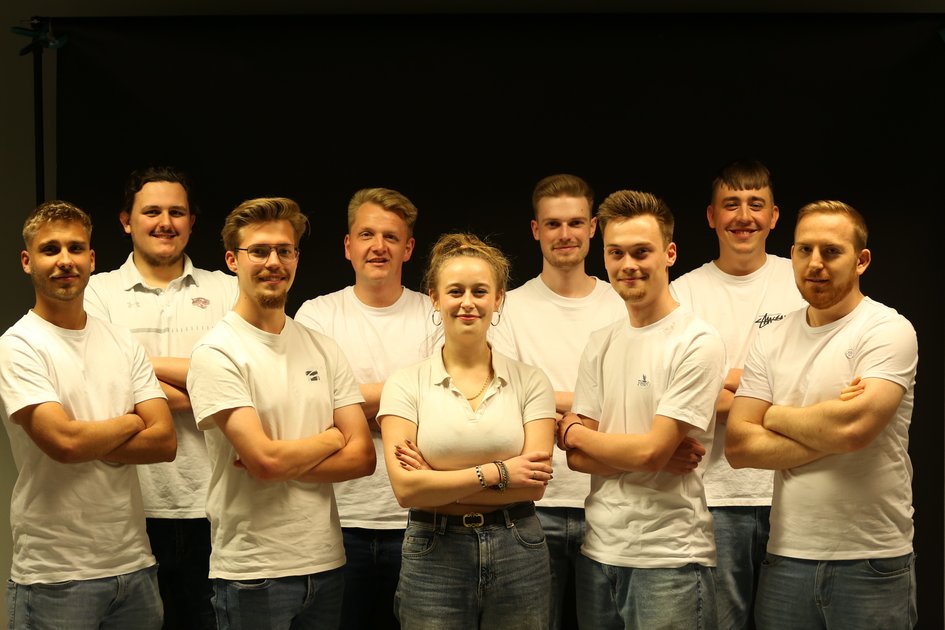 Eine Gruppe von Studierenden stehen in weißen Oberteilen und mit verschränkten Armen vor einem dunklem Hintergrund