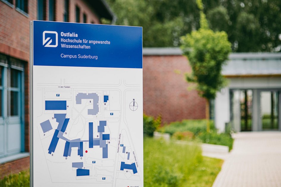 Informationsschild der Ostfalia Hochschule für angewandte Wissenschaften am Campus Suderburg mit Campusplan im Vordergrund; im Hintergrund ist ein moderner Gebäudeeingang sowie Grünflächen zu sehen.