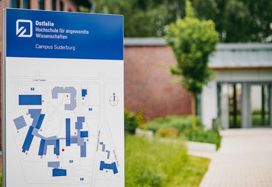 Informationsschild der Ostfalia Hochschule für angewandte Wissenschaften am Campus Suderburg mit Campusplan im Vordergrund; im Hintergrund ist ein moderner Gebäudeeingang sowie Grünflächen zu sehen.