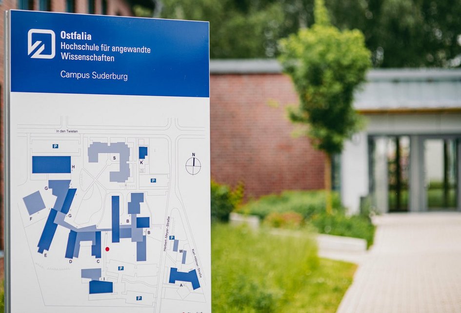 Dekorativ: Informationsschild der Ostfalia Hochschule für angewandte Wissenschaften am Campus Suderburg mit Campusplan im Vordergrund; im Hintergrund ist ein moderner Gebäudeeingang sowie Grünflächen zu sehen.
