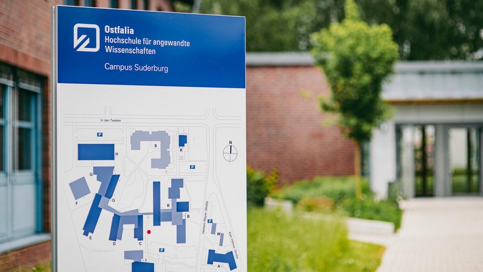 Informationsschild der Ostfalia Hochschule für angewandte Wissenschaften am Campus Suderburg mit Campusplan im Vordergrund; im Hintergrund ist ein moderner Gebäudeeingang sowie Grünflächen zu sehen. (öffnet Vergrößerung des Bildes)
