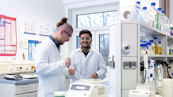 Zwei männlich gelesene Personen stehen nebeneinander in einem Labor