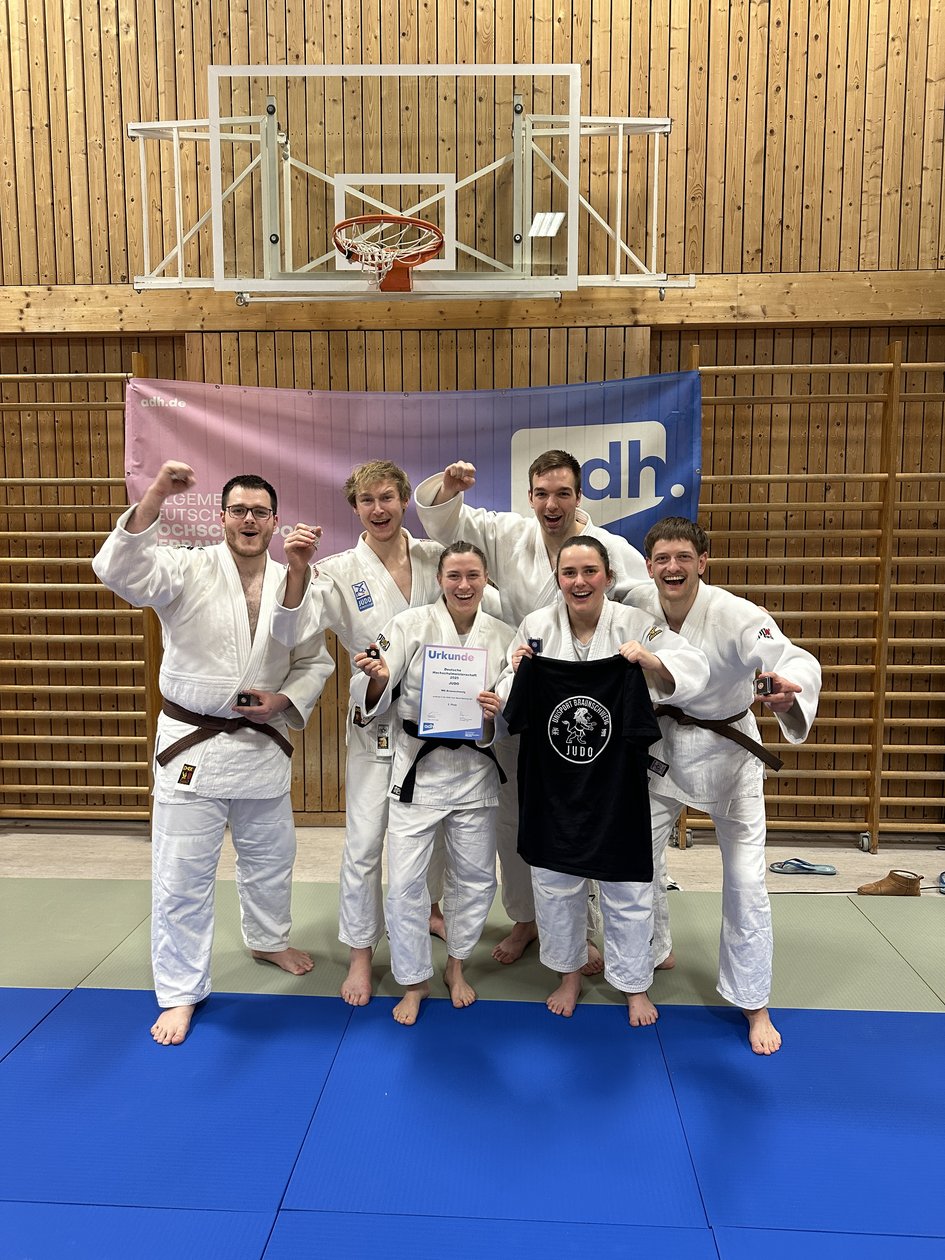 Sechs Judoka jubeln gemeinsam vor einem adh-Banner, präsentieren ihre Bronzemedaillen und die Urkunde der Deutschen Hochschulmeisterschaft im Judo.