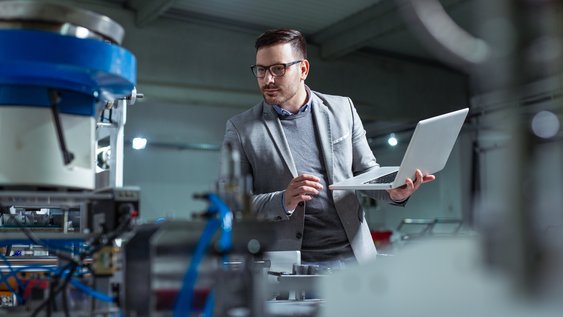 Ein Mann hält seinen Laptop in einer Hand und schaut prüfend auf eine Maschine