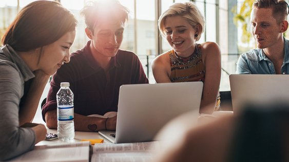 Vier Studierende sitzen vor zwei Laptops, sind gut gelaunt und lernen zusammen.