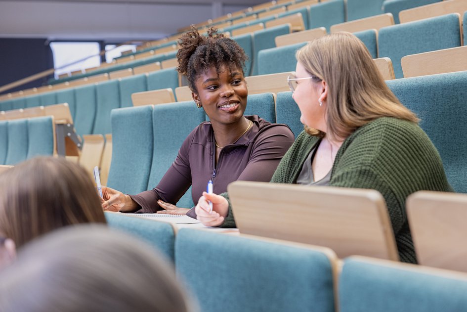 Two female students are sitting in the lecture theatre, chatting 