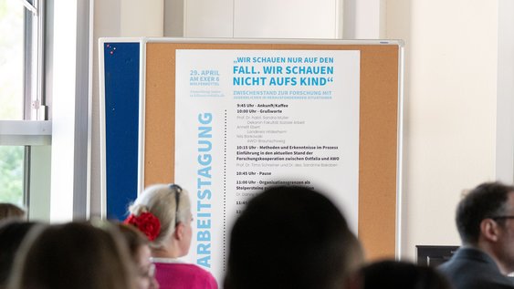 Blick aus Perspektive des Publikums auf das Programm der Tagung, welches vorne links im Saal auf einer Pinnwand angebracht ist.