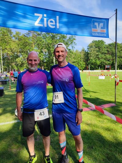 Auf dem Bild sind zwei Teilnehmer des Suderburger Abendlaufs zu sehen, die gemeinsam unter dem Zielbanner der Veranstaltung stehen. Beide tragen die einheitlichen Sporttrikots ihres Teams sowie ihre Startnummern und haben den Lauf erfolgreich beendet. Im Hintergrund ist die grüne Sportanlage mit der Zielstrecke und weiteren Teilnehmern zu erkennen. Die beiden Läufer lächeln zufrieden in die Kamera und zeigen die Freude über ihre Teilnahme an diesem sportlichen Ereignis.