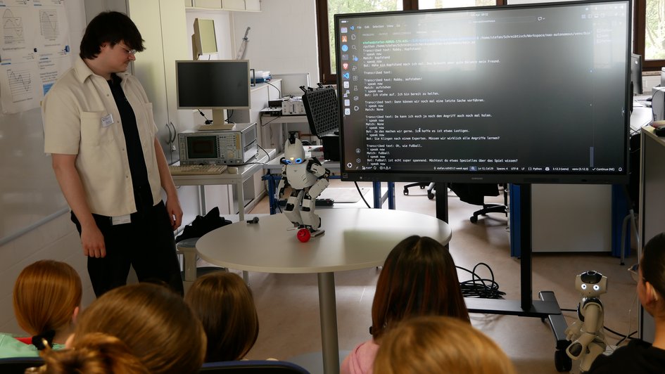 Schülerinnen schauen einem Roboter dabei zu, wie er einen Ball vor sich tritt (öffnet Vergrößerung des Bildes)