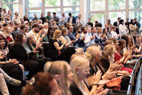 Das Publikum in der Aula klatscht begeistert, während auf der Bühne musiziert wird. (öffnet Vergrößerung des Bildes)
