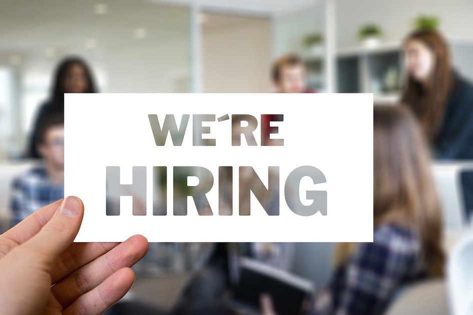 Dekorativ: Hand hält ein Schild mit dem Text 'WE'RE HIRING' vor einer Gruppe von Menschen in einem Büro, symbolisiert Personalmanagement und Rekrutierung.