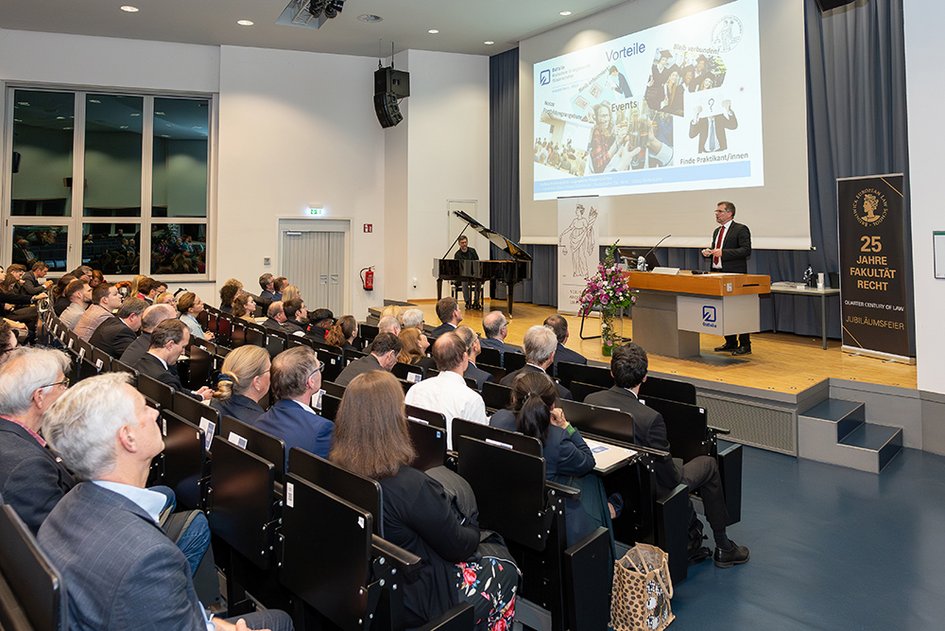 Übersichtsaufnahme mit Blick auf die Bühne, im Vordergrund das Auditorium, am Pult rechts steht Prof. Dr. Litschen, Der Klavierspieler sitzt am Flügel, der links auf der Bühne steht.