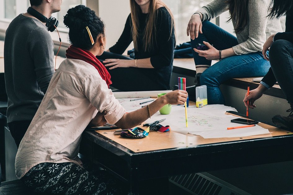 Dekorativ: Vier junge Erwachsene diskutieren und arbeiten gemeinsam an einem Tisch mit Papieren und Stiften, symbolisiert Zusammenarbeit im Bereich Betriebswirtschaft und Management.
