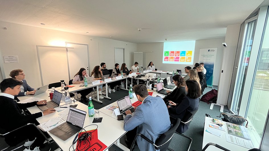 The participants sit at tables arranged in a square and have notebooks in front of them. At the front of the meeting room, 10 SDGs are projected onto the wall via a projector.