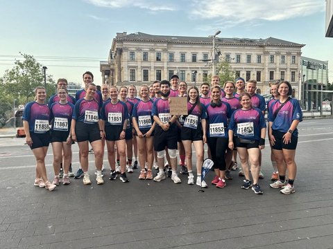 Auf dem Bild ist eine große Gruppe von Läuferinnen und Läufern zu sehen, die gemeinsam für ein Foto posieren. Alle tragen einheitliche Sportkleidung und einige haben ihre Startnummern für den Wettkampf befestigt. Die Gruppe steht vor einem Gebäude in einer Stadt und scheint kurz vor oder nach einer Laufveranstaltung zusammengekommen zu sein. Die vielen Teilnehmer und die gemeinsame Kleidung vermitteln ein Gefühl von Zusammenhalt und Gemeinschaft im Sport.