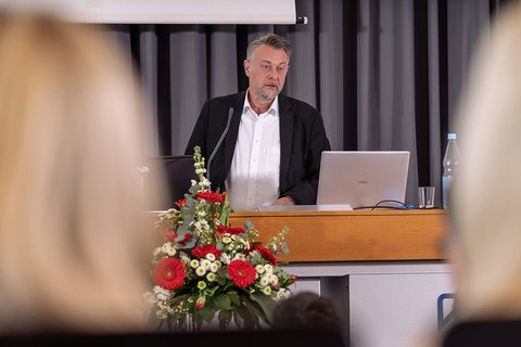 Bürgermeister Ivica Lukanic hält die Laudatio für den Fakultätspreis. Er steht am Pult auf der Bühne, vor dem Pult ist ein Teil des Blumengestecks zu sehen. (öffnet Vergrößerung des Bildes)