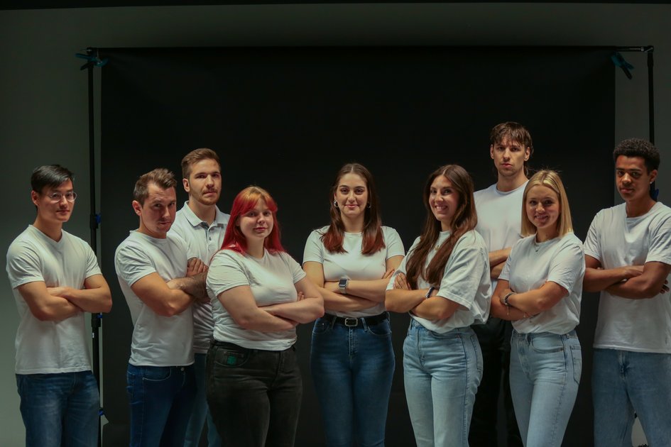Eine Gruppe von Studierenden in weißen Oberteilen stehen mit verschränkten Armen vor einem dunklen Hintergrund