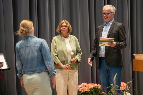 Dekanin Prof. Dr. Sandra Verena Müller hält eine Rose bereit, während Prof. Dr. Georg Kortendieck einer Absolventin die Urkunde überreicht. (öffnet Vergrößerung des Bildes)