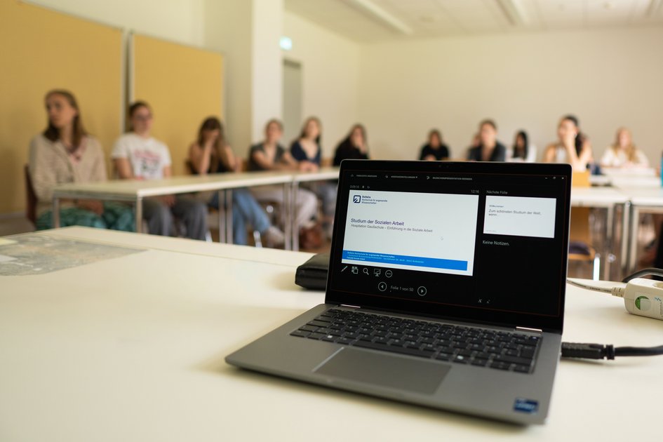 Ein Laptop auf einem Tisch auf dem eine Powerpoint-Präsentation zu sehen ist mit dem Titel "Studium der Sozialen Arbeit". Im Hintergrund sind unscharf Schüler*innen zu sehen, die an Tischen im Seminarraum sitzen.