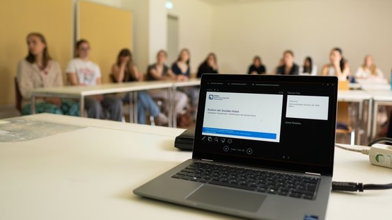 Ein Laptop auf einem Tisch auf dem eine Powerpoint-Präsentation zu sehen ist mit dem Titel "Studium der Sozialen Arbeit". Im Hintergrund sind unscharf Schüler*innen zu sehen, die an Tischen im Seminarraum sitzen.