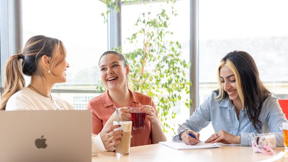 Drei weiblich gelesene Studierende sitzen beinander an einem Tisch, trinken Kaffee und Tee und Lachen.