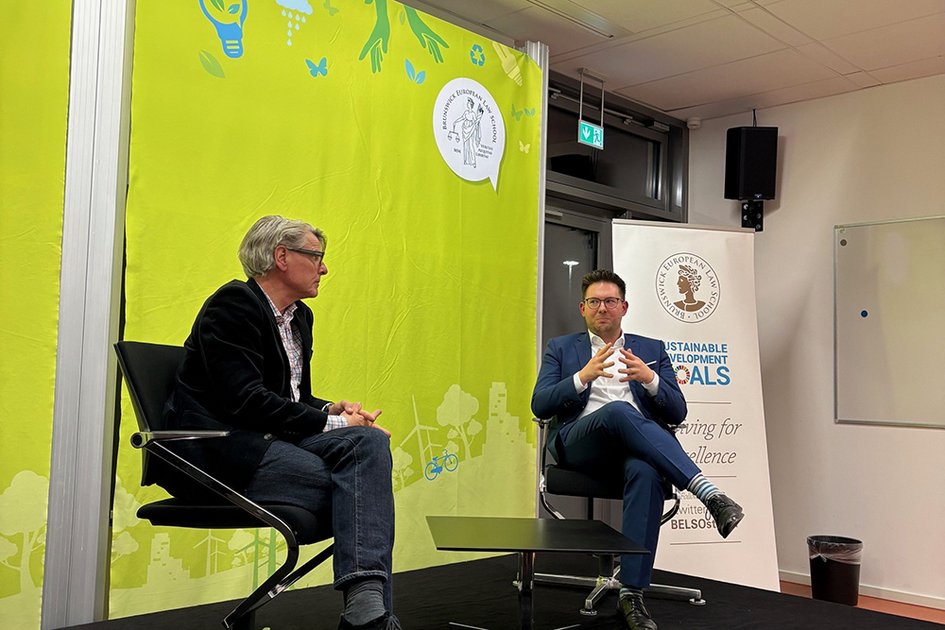 Zwei Personen jeweils in Sakko und Hemd sitzen auf Stühlen auf einem Podium und sind mit einander im Gespräch.