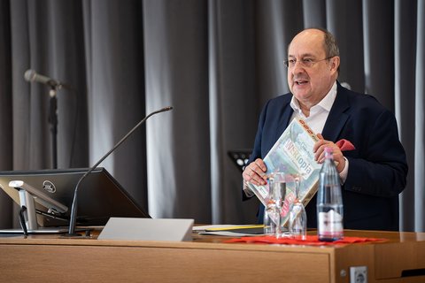 Maic Lindner, stellvertretender Vorstandsvorsitzender der AWO Braunschweig, steht auf der Bühne am Redepult und hält das Spiel "Wertopia" in seinen Händen. (öffnet Vergrößerung des Bildes)