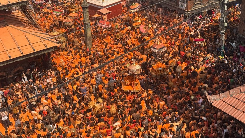 Viele Menschen dichtgedrängt auf einem Platz in Nepal, die Farbe orange dominiert die Bekleidung. Auch im Gesicht haben viele orangene Farbe. Einige runde, schirmartige Gegenstände sind auf dem Platz zu sehen.