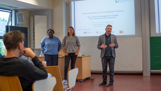 Prof. Dr. Timo Schreiner, Sarah Kilian und Dr. Sandrine Bakoben stehen vorne im Vorlesungssaal und begrüßen alle Anwesenden zu Beginn der Tagung