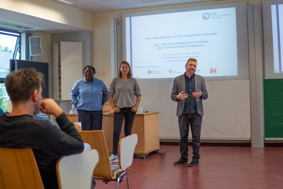 Prof. Dr. Timo Schreiner, Sarah Kilian und Dr. Sandrine Bakoben stehen vorne im Vorlesungssaal und begrüßen alle Anwesenden zu Beginn der Tagung