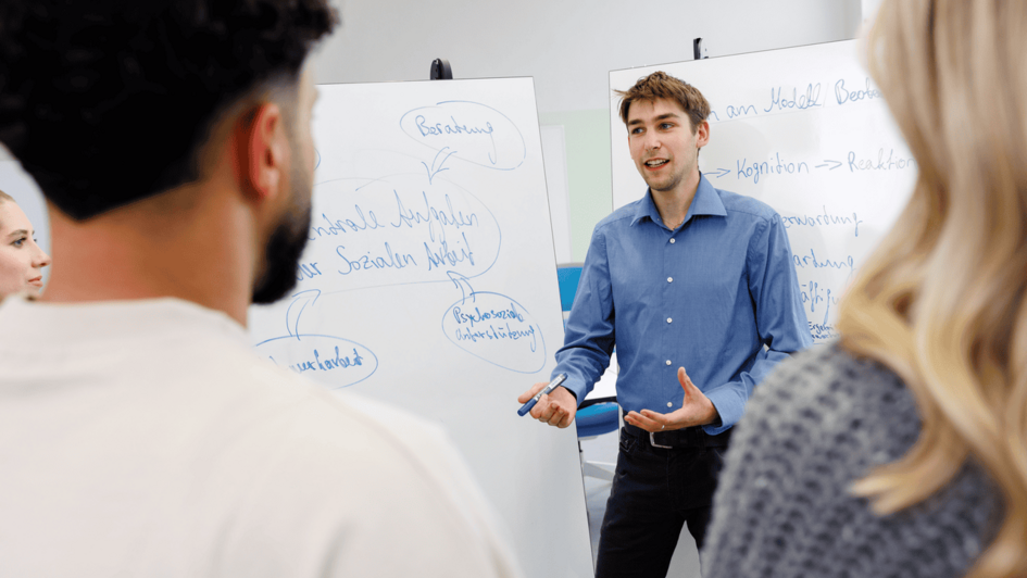 Dekorativ:Studierender in blauem Hemd erklärt vor Flipchart mit handschriftlichen Notizen zwei Personen im Vordergrund. (öffnet Vergrößerung des Bildes)