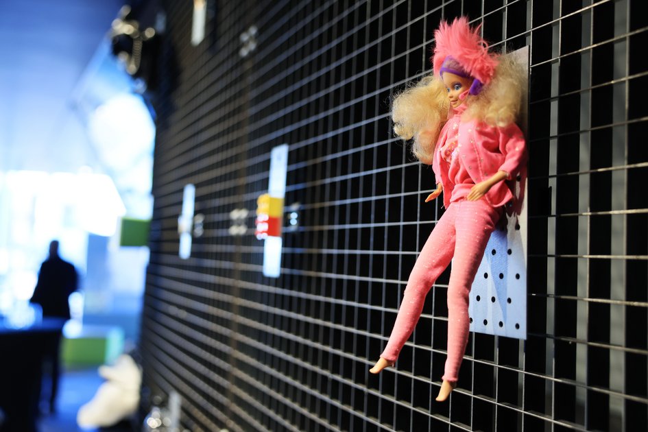 Kunststoffpuppe mit pinker Kleidung und auffälligem pinkem Haarteil an einem schwarzen Ausstellungsgitter.