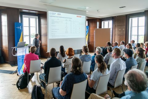 Foto einer Präsentation; vorne wird referiert, während in mehreren Reihen Zuhörer*innen sitzen.