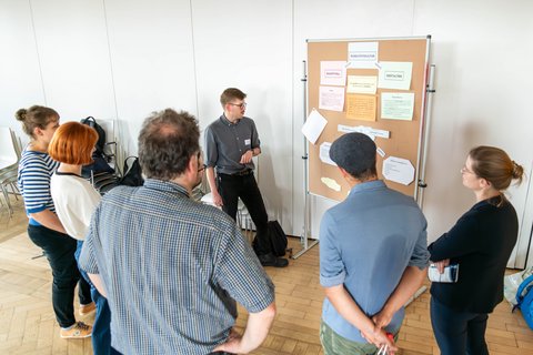 Foto einer Postersession: Eine Gruppe steht vor einer Stellwand auf der ein Plakat zu sehen ist. 