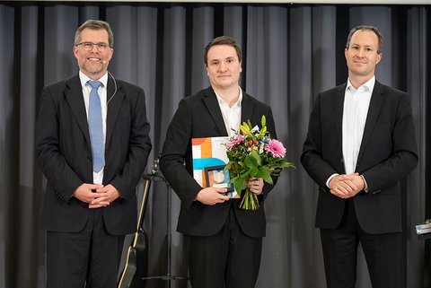 Prof. Dr. Litschen (links), der Preisträger des PKF Fasselt Awards 2025, Dené Schunk und Sören Damerau von PKF Fasselt. Der Preisträger im schwarzen Anzug und weißem Hemd hält seine Auszeichnung und einen bunten Blumenstrauß. (öffnet Vergrößerung des Bildes)