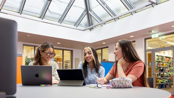 Drei Studentinnen sitzen in der Bibliothek vor Laptops. Sie schauen gemeinsam auf den Bildschirm und lachen.