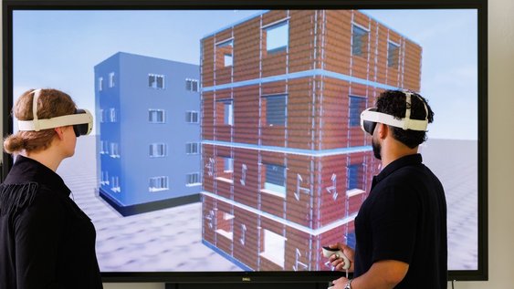 Zwei Studierende mit VR-Brillen stehen vor einem Monitor, auf dem das Gebäudemodell zu sehen ist, das in ihren Brillen projiziert wird.
