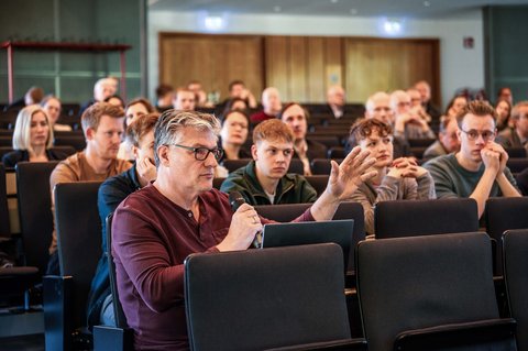 Aufnahme von Zuschauer*innen in der Aula der Ostfalia Hochschule. Ganz vorne hält ein Zuschauer das Mikrofon in der Hand und spricht hinein, während die Zuschauer*innen im Hintergrund gebannt zuhören. (öffnet Vergrößerung des Bildes)